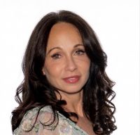 A woman with long, wavy dark hair wearing a light-colored, floral-patterned top looks at the camera against a plain white background.