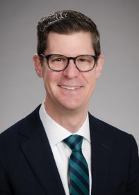 Dr. Jeff Friedrich, MD, wearing glasses, a dark suit, a white shirt, and a green striped tie, smiles in a professional studio portrait with a neutral gray background.