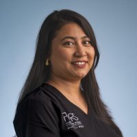 A woman with long dark hair smiles at the camera, wearing a black V-neck shirt with white text on the front, representing Women in Reconstructive Surgery, against a plain light blue background.
