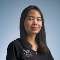 A woman with long black hair, wearing a black top with white writing and a gold necklace, poses in front of a plain light blue background, smiling gently at the camera—an inspiring presence among Women in Reconstructive Surgery.