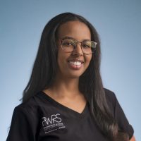 A woman with long dark hair and glasses smiles at the camera. She is wearing a black V-neck "PWRS Pioneering Women" top, representing Women in Reconstructive Surgery, and stands in front of a plain blue background.