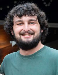 A young man with curly dark hair and a full beard is smiling at the camera. Micah Warner-Carey is wearing a green t-shirt and standing in an outdoor setting with blurred lights in the background.