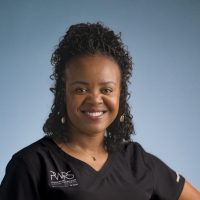 A woman with curly hair, representing Women in Reconstructive Surgery, smiles confidently in a black shirt with a "PMRS" logo against a plain blue background.