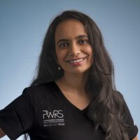 A woman with long dark hair smiles at the camera, wearing a black shirt featuring the Women in Reconstructive Surgery logo, standing against a plain blue background.