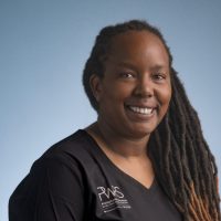 A woman with long, dark, and light-tipped locs smiles at the camera. She wears a black V-neck shirt with a logo and text reading "RMS," representing Women in Reconstructive Surgery, and stands against a plain blue background.